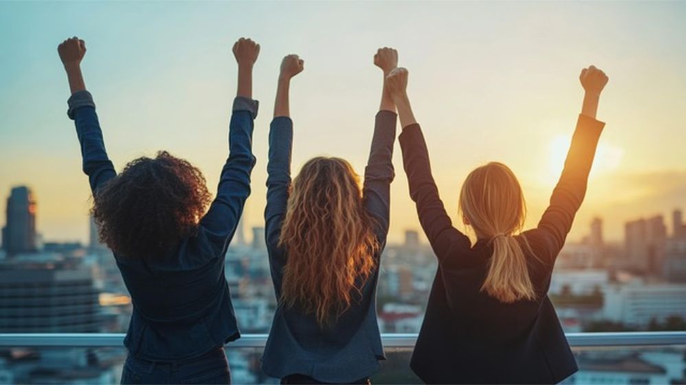 Three women stand in the sunrise, their backs to the viewer. Their fists are raised over their heads. Victory!